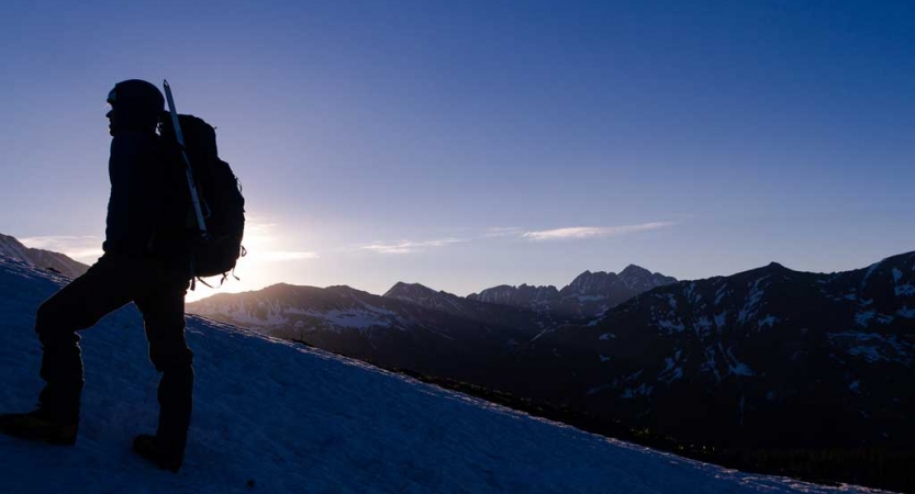 The silhouette of a person wearing a backpack is illuminated by the sun behind them. They appear to be standing on a snowy incline with mountains in the background.
